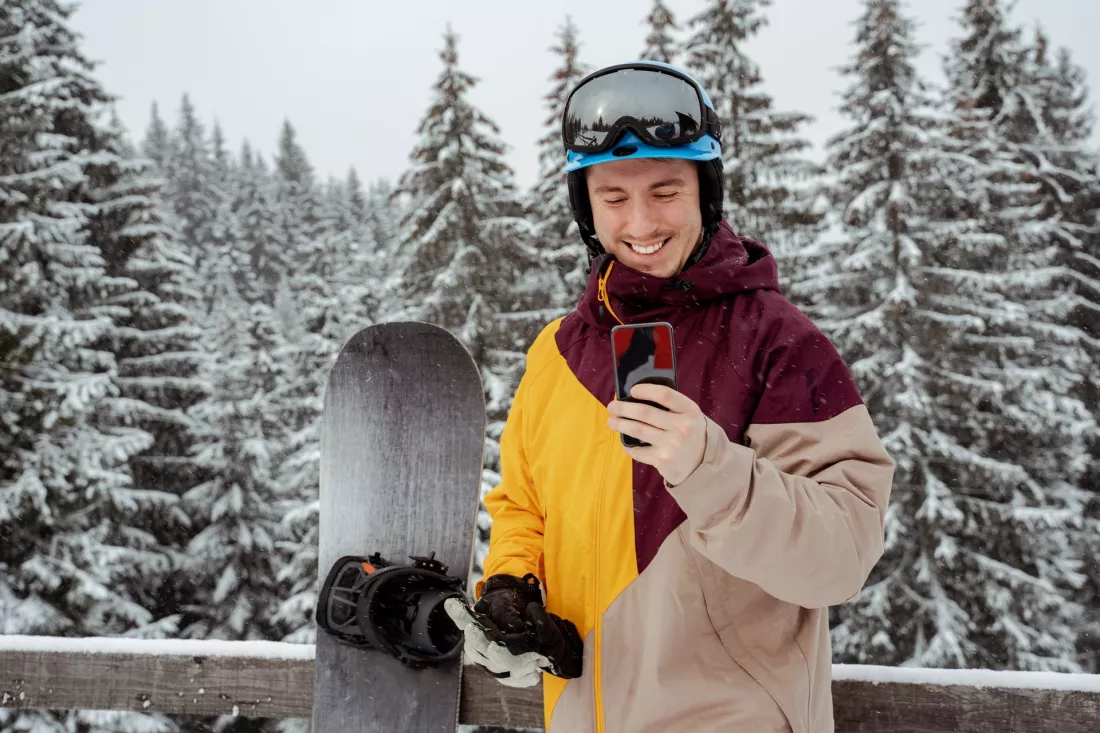 Snowboardzista w kasku i goglach patrzy na telefon na tle ośnieżonego lasu