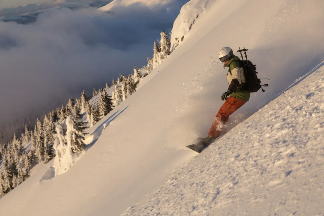 Snowboardzista z plecakiem i kaskiem zjeżdża stromym, nieprzetartym stokiem w głębokim puchu, poniżej morze chmur.
